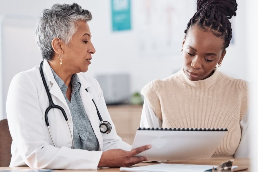 A imagem mostra duas mulheres em um ambiente que aparenta ser um consultório médico. À esquerda, uma profissional de saúde, usando jaleco branco e estetoscópio, conversa com a paciente enquanto apresenta informações em um bloco ou documento. A paciente observa atentamente o material durante a orientação.