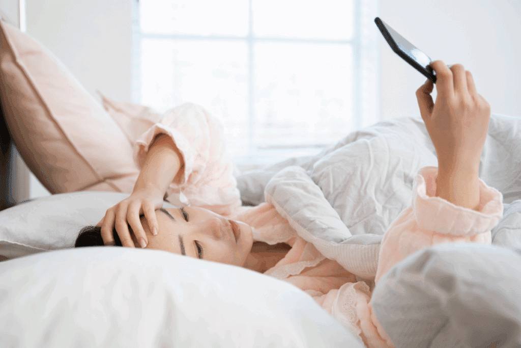 Mulher deitada na cama usando pijama laranja com uma mão na cabeça e a outra com um celular olhando notificações após acordar