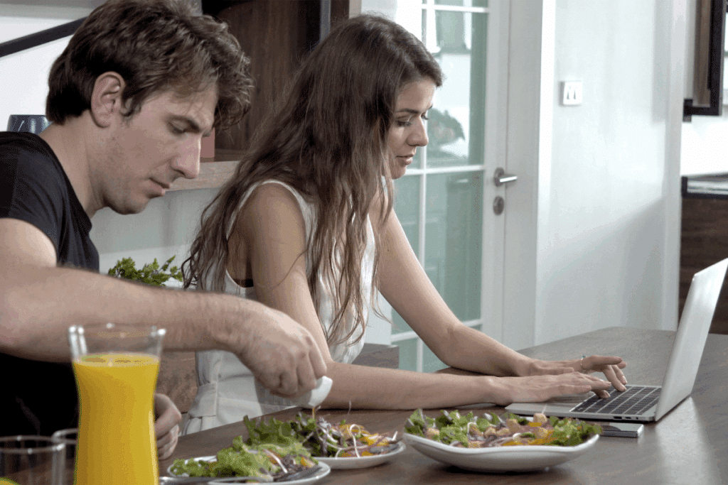 Homem comendo salada e mulher ao lado mexendo em um notebook