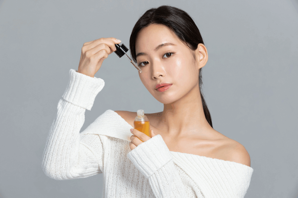 A imagem mostra uma mulher segurando um frasco de produto para cuidados com a pele e um conta-gotas próximo ao rosto. Ela veste uma blusa branca e aparece diante de um fundo cinza neutro. O líquido no recipiente tem tonalidade amarelada e lembra um sérum ou óleo facial.
