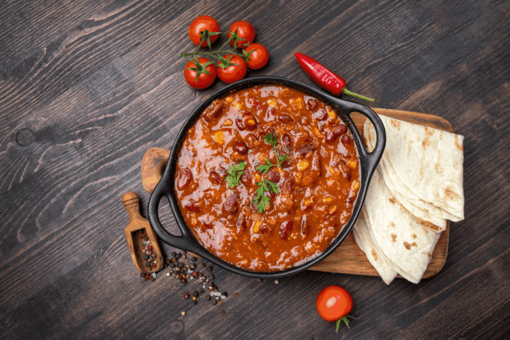 A imagem mostra uma panela de ferro preta cheia de um ensopado espesso à base de feijão vermelho, com molho avermelhado e grãos de milho visíveis, decorado com um ramo de salsa no centro. Ao lado da panela, sobre uma tábua de madeira, há tortilhas dobradas, prontas para acompanhar a refeição.