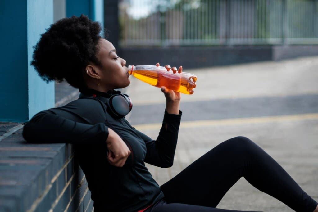 Uma mulher negra usando roupas de ginástica pretas e fones de ouvido no pescoço, sentada e bebendo um líquido laranja de uma garrafa de plástica