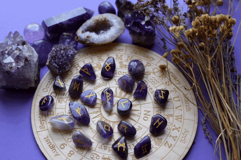 A imagem apresenta uma composição temática voltada ao universo do esoterismo e das práticas divinatórias. Em destaque, há diversas pedras de ametista gravadas com símbolos rúnicos dourados, organizadas sobre um tabuleiro circular decorado com referências ao zodíaco, aos meses do ano e a elementos astrológicos.