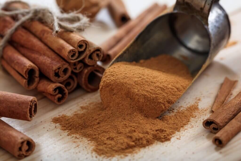 Canela em pó sendo despejada de uma colher metálica ao lado de paus de canela sobre superfície de madeira
