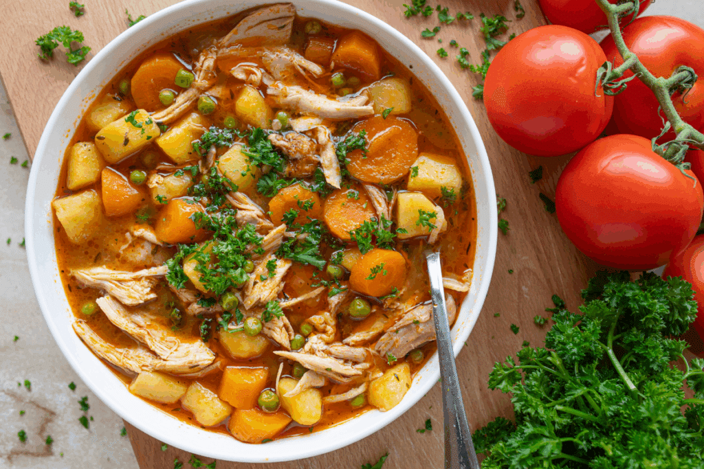 Sopa de frango com batata, cenoura e ervilha servida em tigela branca ao lado de tomate e ramo de salsa em cima de mesa de madeira