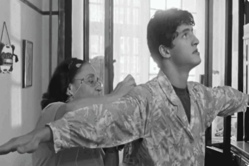 Imagem em preto e branco de mulher idosa fazendo ajuste em roupa de um jovem com cabelo curto usando camisa estampada