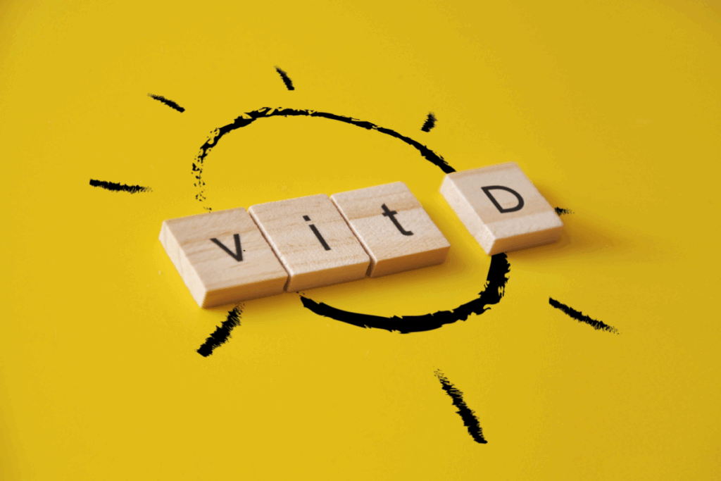 Blocos de madeira formam a palavra "Vit D" sobre um fundo amarelo, acompanhados da ilustração de um sol, representando a vitamina D e sua relação com a exposição solar