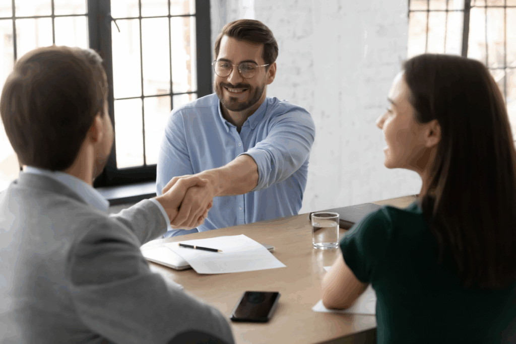 Três pessoas reunidas ao redor de uma mesa de escritório durante um atendimento ou reunião de negócios. No centro, um homem sorridente de camisa azul e óculos cumprimenta o cliente com um aperto de mão por cima da mesa. Ao lado, uma mulher observa a interação, enquanto papéis e uma caneta estão posicionados sobre a mesa.