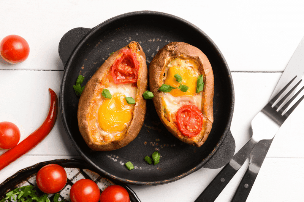 Duas metades de batata-doce assada recheadas com ovo frito de gema mole e fatia de tomate, salpicadas com cebolinha picada e servidas em uma frigideira de ferro preta.