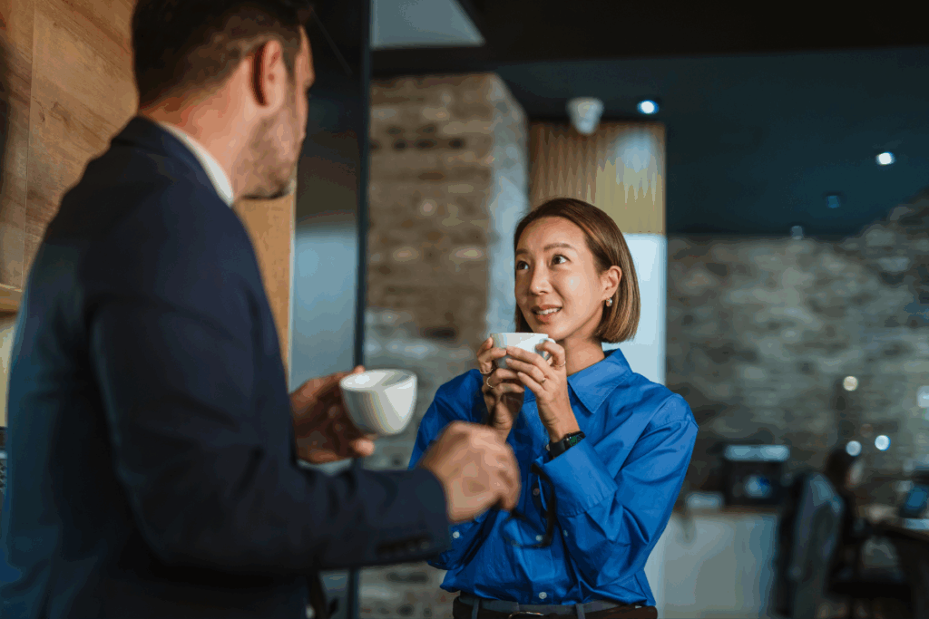Uma mulher de camisa azul-brilhante segura uma xícara branca perto do rosto e conversa sorrindo com um colega de terno escuro, visto de costas, em uma área de café moderna.