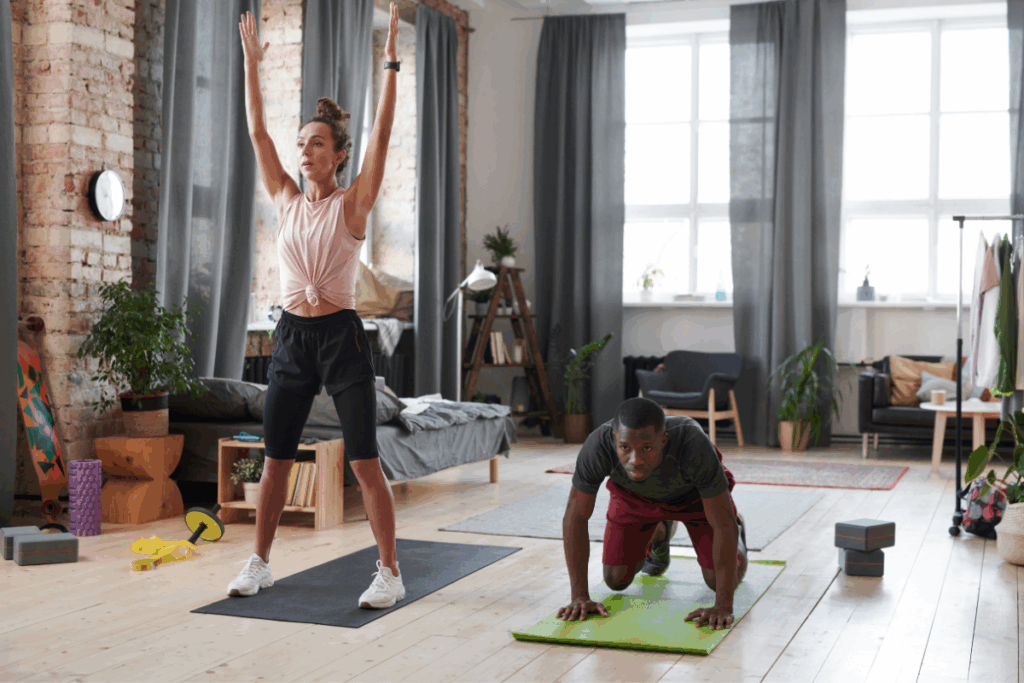 Homem e mulher praticando exercícios físicos juntos sobre colchonetes em uma sala ampla. Em primeiro plano, a mulher está de pé com os braços levantados para o alto, enquanto o homem, ao lado, está agachado apoiando as mãos no chão.