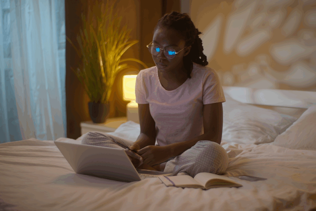 Mulher negra de óculos e pijama sentada de pernas cruzadas em uma cama, olhando para a tela de um notebook aberto, com um caderno ao seu lado.