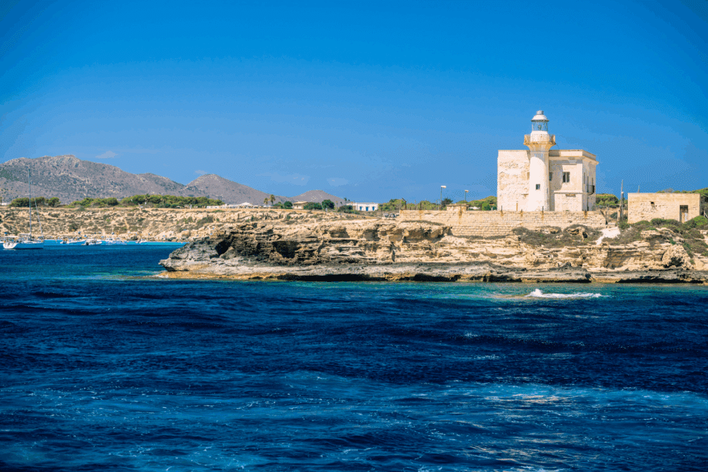 Farol histórico de alvenaria clara sobre promontório rochoso à beira-mar, sob céu azul límpido. Em primeiro plano, mar em tons intensos de azul e turquesa; ao fundo, relevo montanhoso e pequenas embarcações na costa