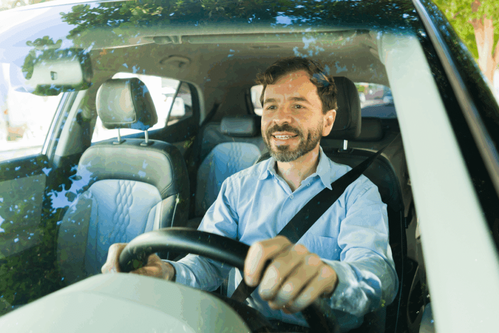Um homem de meia-idade, com barba e vestindo uma camisa de botão azul claro, está sorrindo enquanto segura o volante e olha para a frente. Ele está usando o cinto de segurança. A foto é tirada de fora, olhando através do para-brisas, que reflete as árvores próximas.