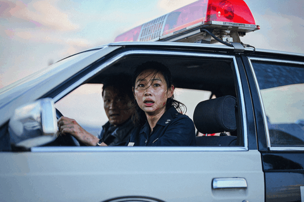 Uma mulher de uniforme policial com marcas no rosto olhando aflita pela janela de um carro de polícia com giroflex vermelho, enquanto um homem dirige ao seu lado.