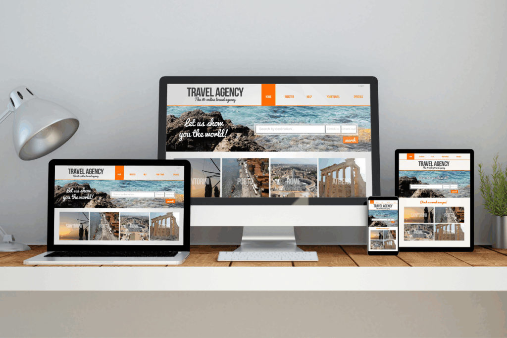 Interface de um site de agência de viagens adaptada para múltiplos dispositivos, um monitor desktop central, um notebook à esquerda, um tablet à direita e um smartphone à frente, todos dispostos sobre uma mesa de madeira ao lado de uma luminária metálica e um pequeno vaso de planta. O layout do site traz um cabeçalho laranja e branco, barras de busca de destinos e fotos em miniatura destacando destinos turísticos como Santorini, Porto, Roma e Atenas