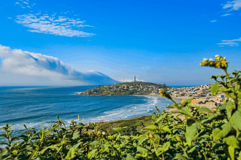 Vista panorâmica do litoral de Laguna (SC), destacando a praia com vegetação em primeiro plano e o promontório com o icônico Farol de Santa Marta ao fundo