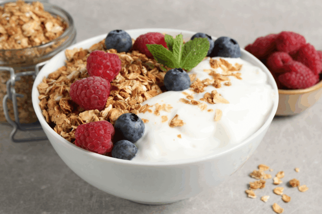 Tigela branca servida com iogurte natural, granola crocante e frutas vermelhas frescas, como framboesas e mirtilos, decorada com um ramo de hortelã no topo, além de potes ao fundo com mais granola e framboesas