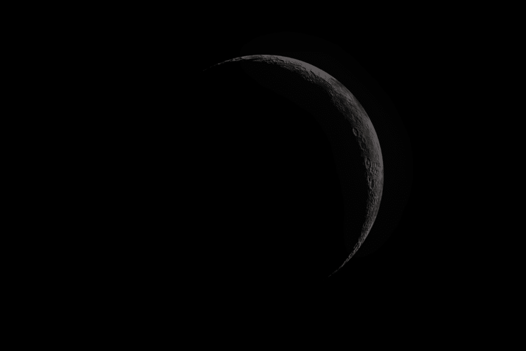 Uma imagem de uma fina lua minguante iluminada no céu noturno profundo, isolada contra um fundo preto. Apenas uma pequena porção da lua é visível como uma faixa estreita e brilhante no lado direito, revelando craters e texturas de superfície detalhadas.
