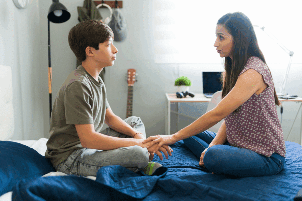 Uma mulher e um menino adolescente estão sentados de frente um para o outro em uma cama com colcha azul. Ela segura a mão dele carinhosamente enquanto conversa com uma expressão séria e acolhedora.