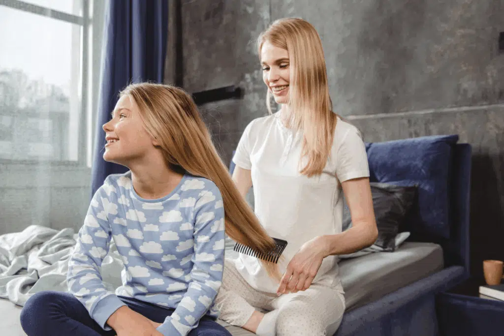 Mulher loira sorridente penteia o cabelo longo e loiro de uma menina sentada à sua frente na cama. A menina veste um pijama azul com estampa de nuvens e olha para o lado sorrindo.