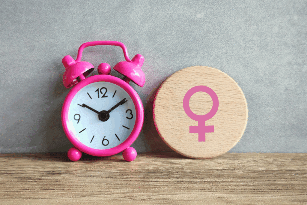 Um relógio despertador rosa pequeno posicionado ao lado de um disco de madeira que exibe o símbolo do gênero feminino estampado em rosa, ambos sobre uma superfície de madeira diante de uma parede cinza.
