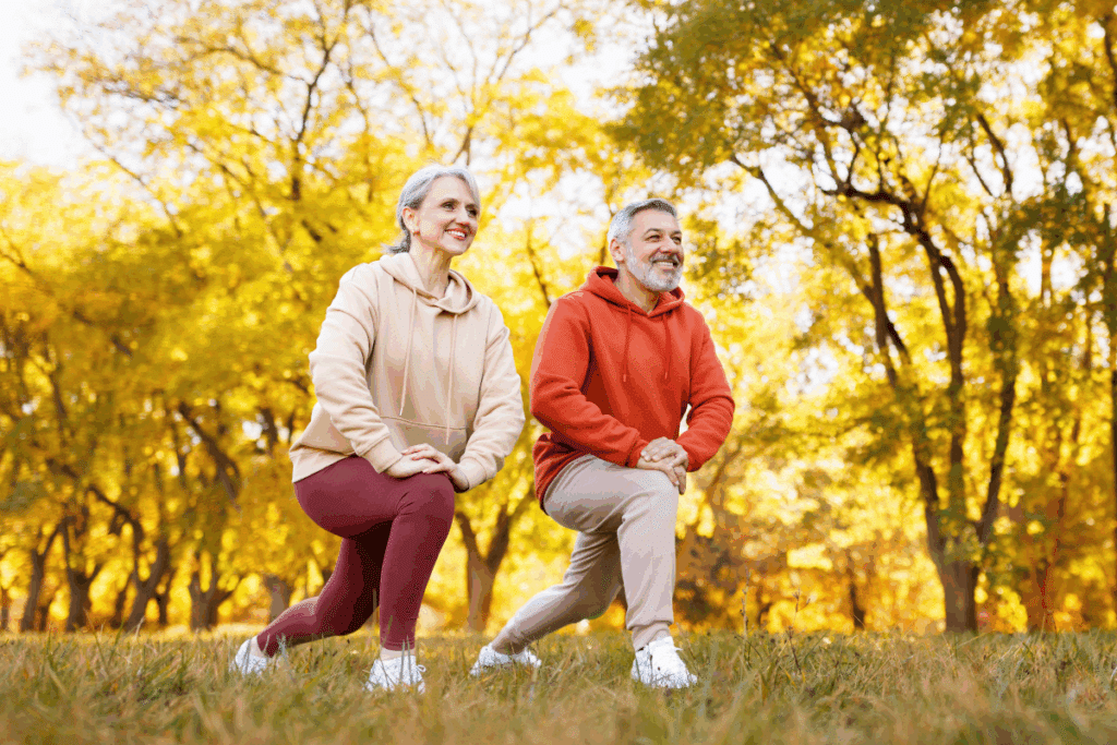 Casal de idosos sorridentes ao ar livre fazendo exercício de afundo (alongamento de pernas) em um parque durante o outono, cercados por árvores com folhas amareladas.