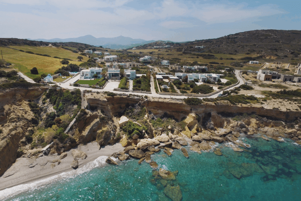 Vista aérea de uma costa litorânea com falésias rochosas e construções de arquitetura cicládica branca. Na base da rocha, uma pequena faixa de praia e águas cristalinas em tons de turquesa, com colinas suaves e vegetação baixa ao fundo