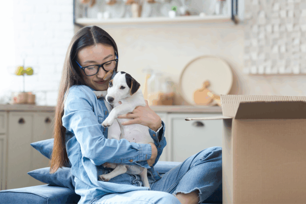 Uma mulher asiática com longos cabelos escuros e óculos, vestindo uma camisa jeans e calça jeans rasgada, sorri e abraça um cachorrinho de cor branca com manchas pretas e marrons. Ela está sentada em um sofá azul e o cachorrinho está sendo segurado confortavelmente em seus braços. Há uma caixa de papelão aberta no canto inferior direito da imagem.