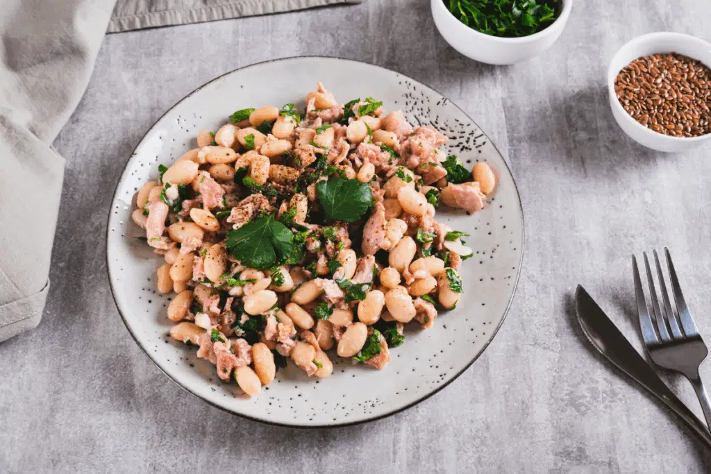 Prato raso e claro com pintas pretas servindo uma salada de feijão-branco misturado com pedaços de atum e salsa fresca picada, temperada com pimenta-do-reino. O prato está colocado sobre uma superfície cinza neutra, acompanhado por um garfo e uma faca à direita, e pequenas tigelas com temperos ao fundo.