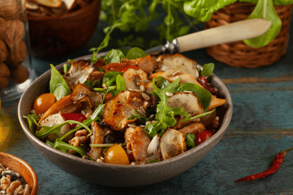 Tigela cinza com salada colorida composta por folhas de rúcula, tomates-cereja amarelos e vermelhos, pedaços de frango grelhado, chips de topinambur (tupinambo) dourados e sementes polvilhadas por cima. Ao fundo, veem-se potes de madeira com nozes e sementes sobre uma mesa rústica.