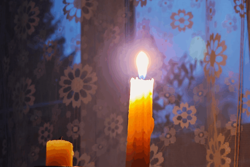 Vela amarela acesa em primeiro plano com uma cortina floral transparente ao fundo, sob luz azulada.