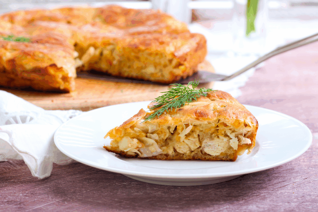 Uma fatia de torta salgada recheada com frango servida em um prato branco raso, decorada com um ramo de endro (dill) no topo. Ao fundo, desfocado, sobressai o restante da torta dourada sobre uma tábua de madeira.