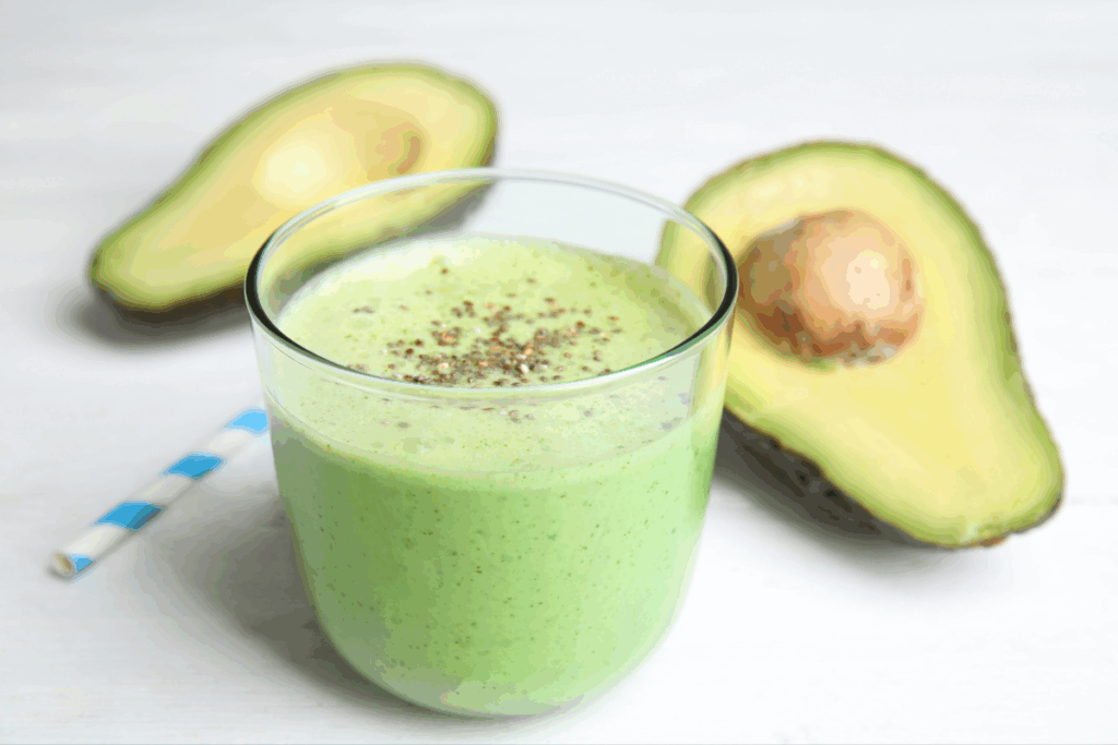 Copo transparente com vitamina verde coberta com sementes de chia, posicionado sobre uma superfície branca ao lado de metades de abacate e um canudo listrado de azul e branco.