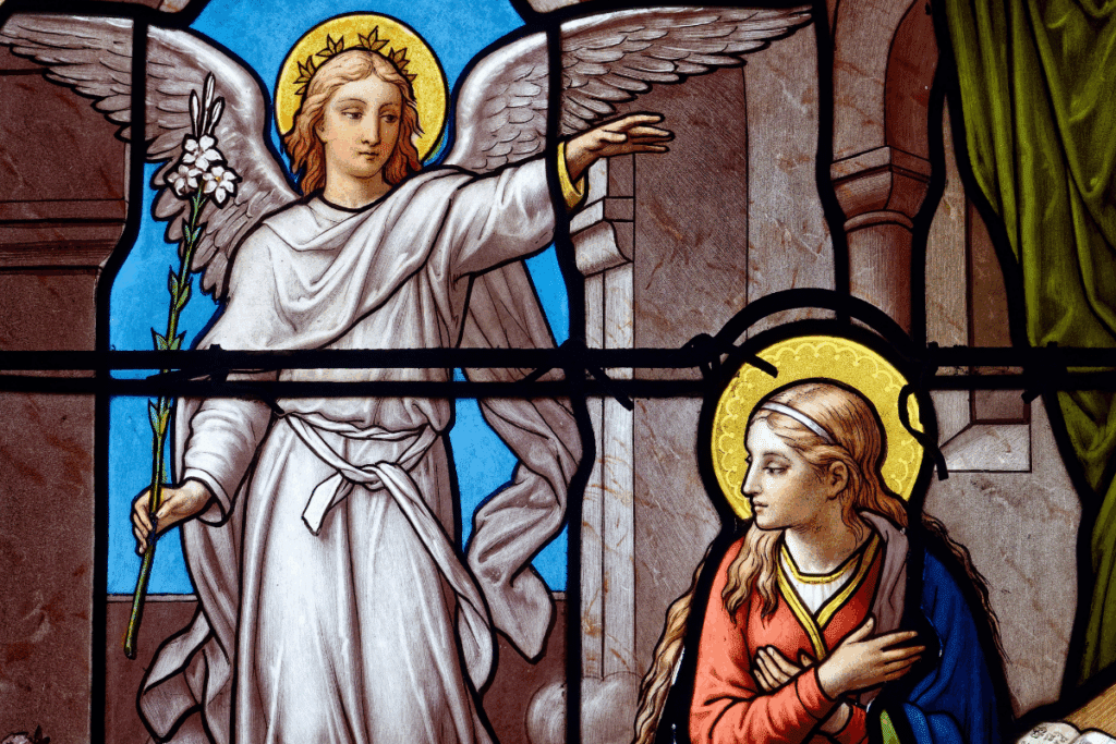 Vitral religioso representando a Anunciação. À esquerda, o Arcanjo Gabriel, em vestes brancas com asas abertas e auréola dourada, segura um ramo de lírio branco e estende a mão em direção à Virgem Maria. À direita, Maria aparece de perfil, vestindo túnica vermelha e manto azul, com a cabeça levemente inclinada e as mãos sobre o peito