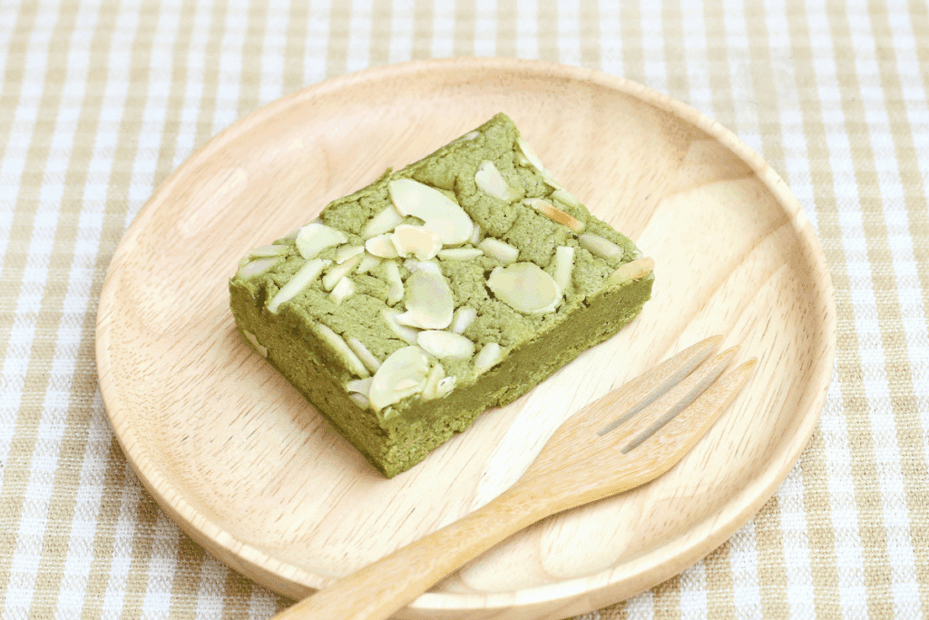 Brownie de matcha cortado em formato quadrado, servido individualmente sobre um prato redondo de madeira clara. O doce tem coloração verde-clara, textura densa e macia e é coberto com lâminas de amêndoas. Ao lado do brownie há um pequeno garfo de madeira clara. O prato está sobre uma toalha de mesa com estampa xadrez em tons de branco e bege, criando uma composição delicada e clara.