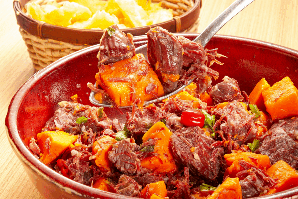Carne de sol desfiada com pedaços de abóbora cozida, servida em uma travessa de cerâmica vermelha sobre uma mesa de madeira.