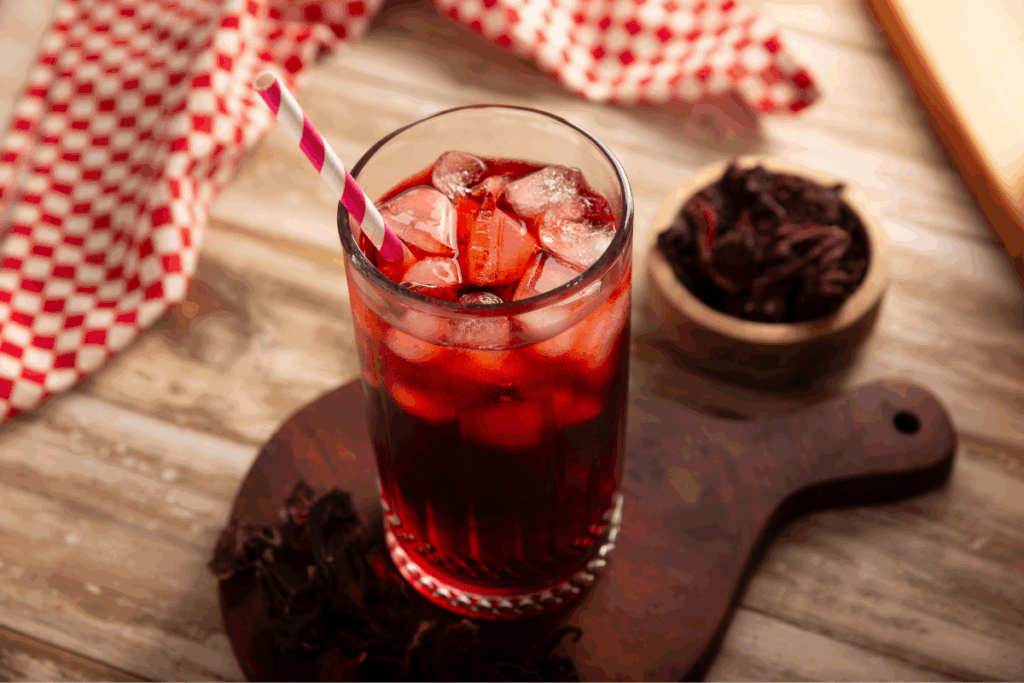 Um copo alto com chá gelado de hibisco, de cor vermelho-intensa, repleto de cubos de gelo e com um canudo listrado em rosa e branco. O copo está apoiado em uma tábua de corte de madeira escura. Ao redor do copo e dentro de um pequeno recipiente de madeira ao lado, veem-se flores secas de hibisco. O cenário é composto por uma mesa de madeira clara e um pano xadrez vermelho e branco ao fundo