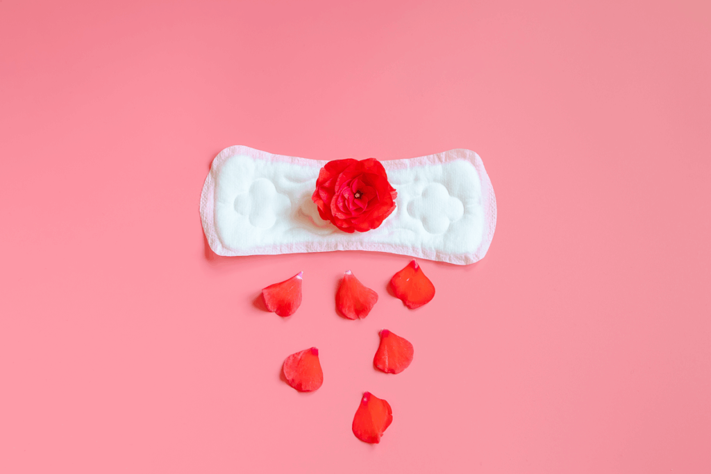 Absorvente higiênico branco com uma rosa vermelha ao centro e pétalas espalhadas como gotas sobre fundo rosa, representando o fluxo menstrual e as cólicas menstruais.