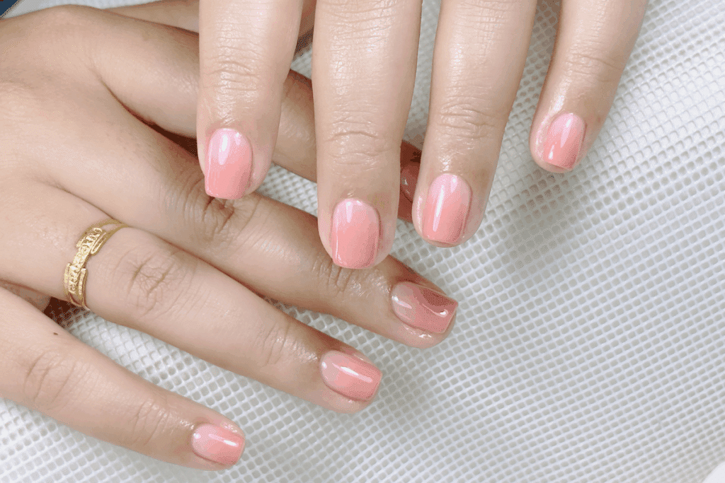 Mãos femininas apoiadas sobre uma superfície rendada branca, exibindo unhas curtas pintadas no estilo syrup nails em tom rosa-goiaba translúcido e acabamento brilhante. Na mão esquerda, há um anel dourado delicado no anelar
