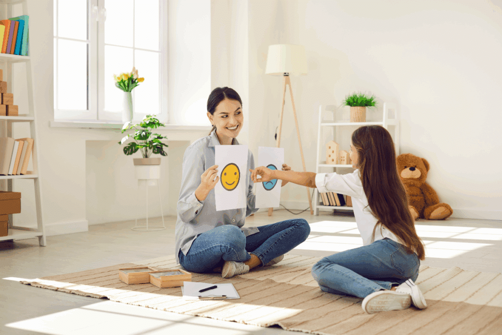 Uma mulher adulta e uma menina sentadas no chão de um ambiente claro. A mulher segura cartões com diferentes expressões faciais, enquanto a menina aponta para um deles. Ao fundo, há estantes, livros, plantas, uma janela e um urso de pelúcia.