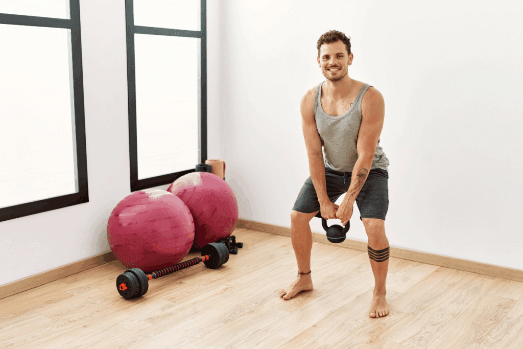 Homem jovem realizando agachamento descalço com kettlebell em uma sala de treino iluminada por luz natural. Veste regata cinza e bermuda escura. Ao fundo, piso amadeirado, duas bolas de pilates na cor rosa-queimado e uma barra com anilhas no chão