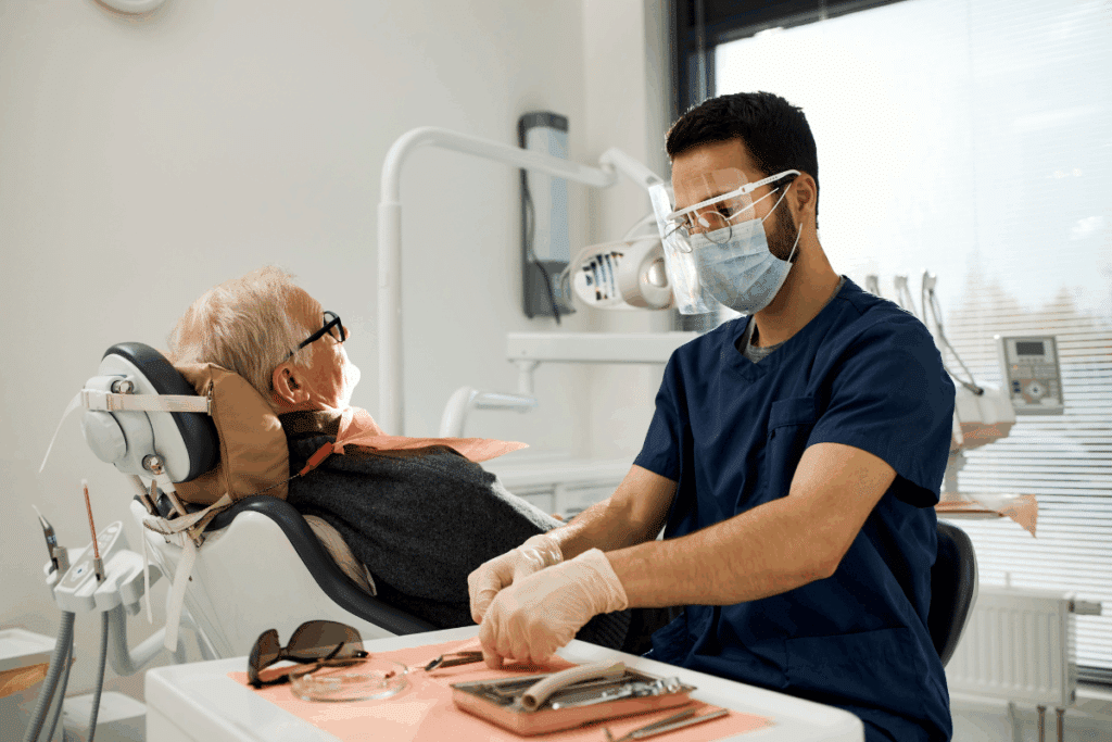 Dentista de máscara e protetor facial organizando seus instrumentos em uma mesa, enquanto um homem idoso aguarda deitado na cadeira odontológica ao fundo