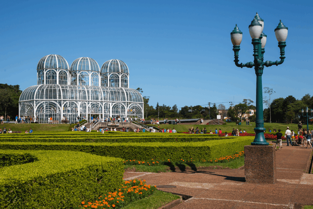 Estufa de vidro e estrutura metálica branca do Jardim Botânico de Curitiba, cercada por jardins geométricos com flores laranjas e visitantes caminhando.
