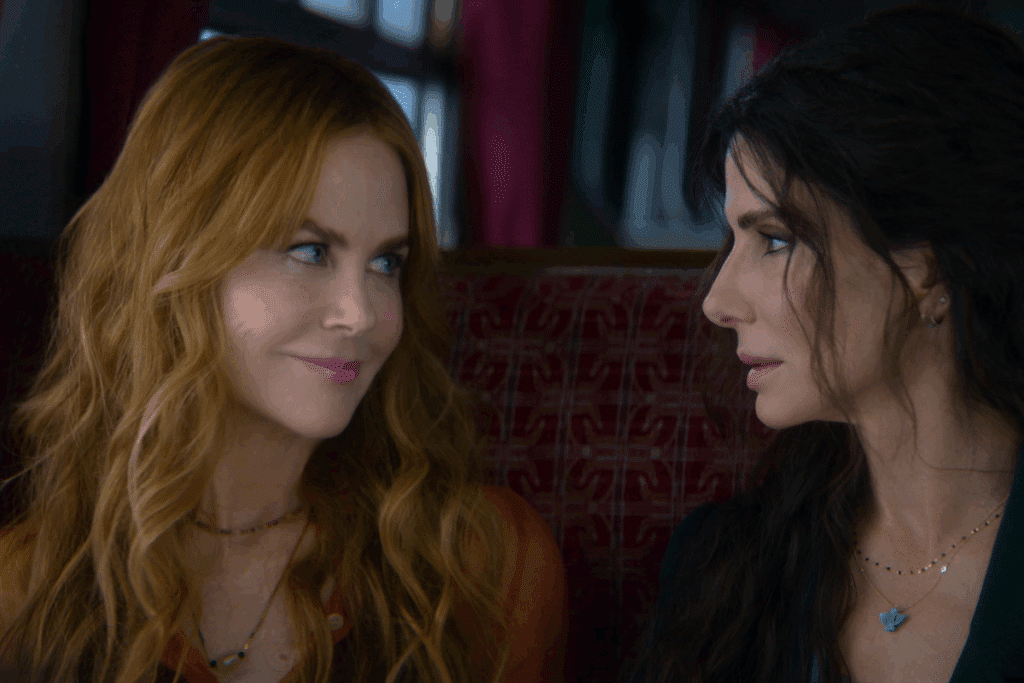 Nicole Kidman e Sandra Bullock como as irmãs bruxas Gillian e Sally, sentadas lado a lado em um banco de estofado vermelho e trocando um olhar cúmplice, em cena de "Da Magia à Sedução: Feitiço de Amor".