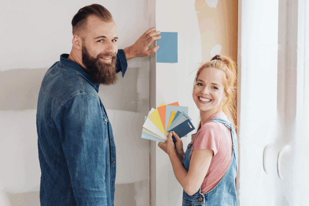 Um casal sorridente trabalhando na remodelação de um ambiente, com o homem a encostar uma amostra de tinta azul à parede enquanto a mulher segura um leque com várias paletas de cores