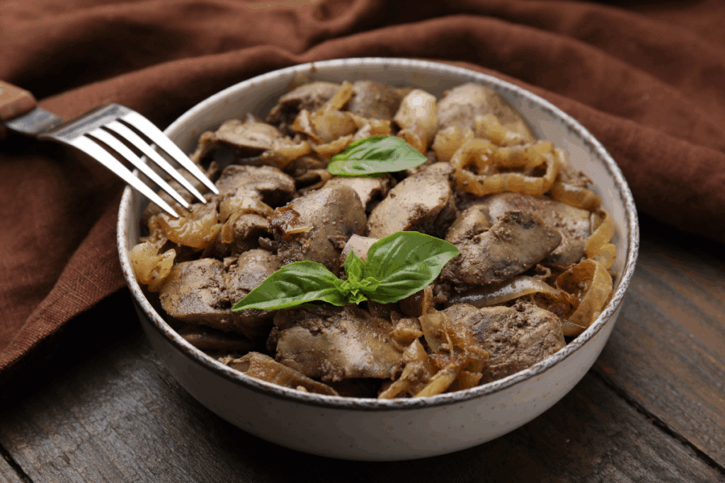 Prato de fígado de frango acebolado servido em uma tigela branca de cerâmica, acompanhado de cebolas douradas e decorado com folhas frescas de manjericão. Um garfo aparece apoiado sobre a lateral do prato. A composição está sobre uma mesa de madeira escura, com um tecido marrom ao fundo, criando um ambiente rústico e aconchegante.