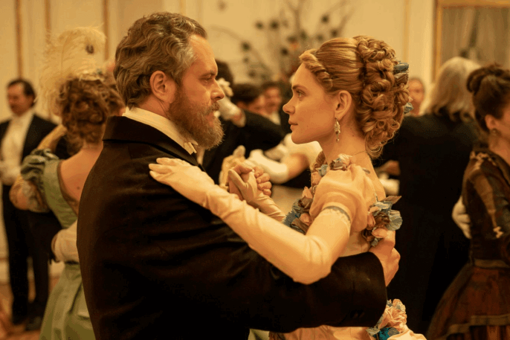 Cena de baile de época, com um casal dançando em meio a outros convidados. A mulher usa um vestido elegante com detalhes florais e luvas longas, enquanto o homem veste um traje social preto. O ambiente é sofisticado e iluminado em tons quentes.