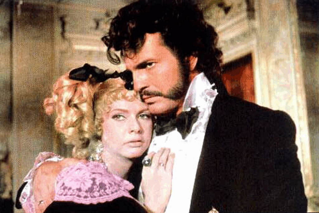 Cena de um filme de época com um homem de cabelos escuros, barba e bigode, usando um traje preto com camisa branca e lenço no pescoço. Ao lado dele, uma mulher de cabelos loiros presos e vestido rosa olha para a frente. Os dois aparecem em um ambiente interno.