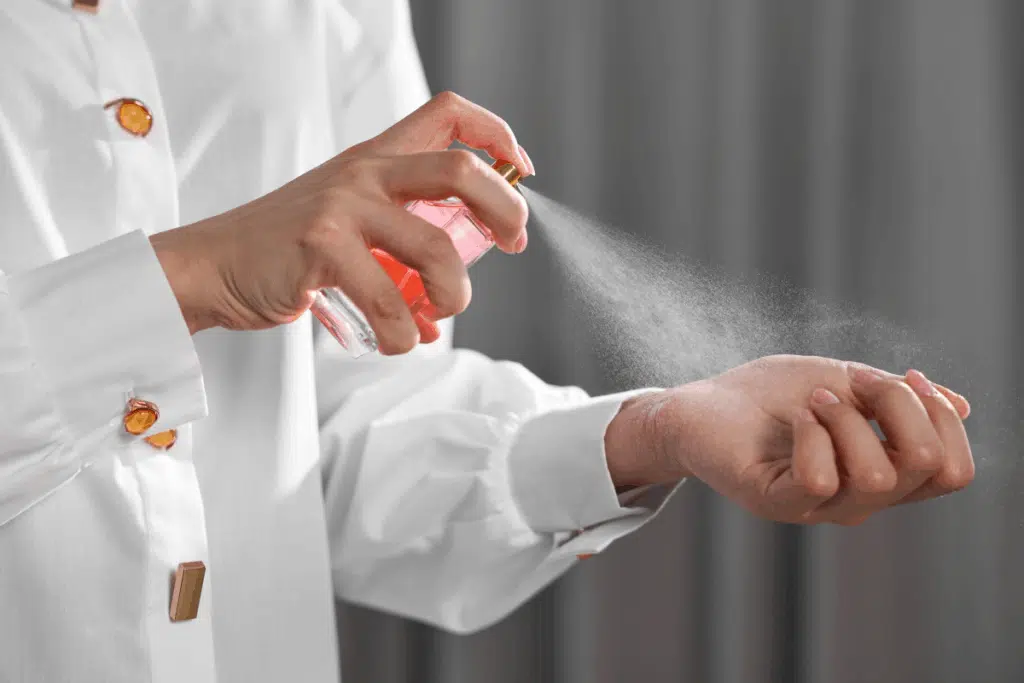 Plano aproximado das mãos de uma pessoa a vestir uma camisa branca elegante com botões dourados. Com a mão direita, segura um frasco de perfume de tom rosa e borrifa uma névoa fina da fragrância diretamente sobre o pulso esquerdo